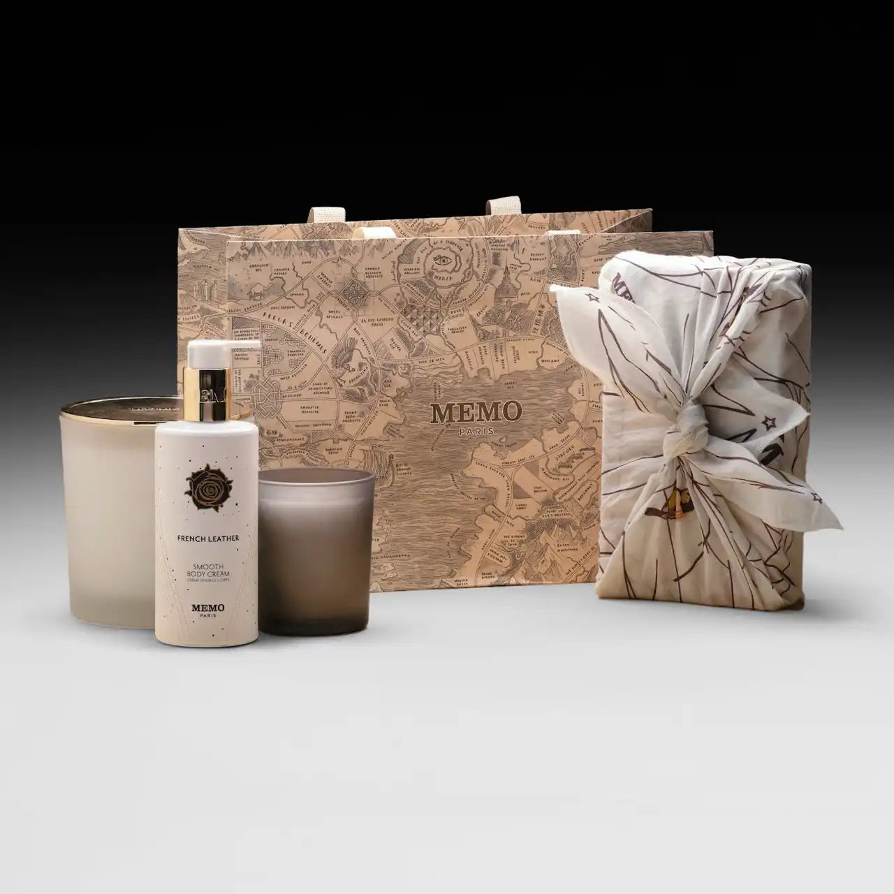 A collection of Memo Paris brand fragrance products, including a bottle of ’French Leather’ perfume, two candles, and a gift bag, presented as a gift set.