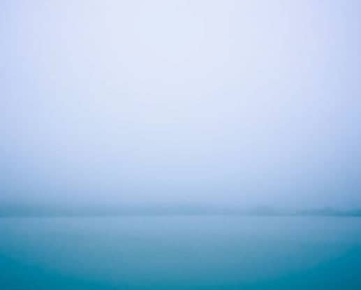 A serene, misty horizon where soft blue tones blend into a hazy sky above calm water.